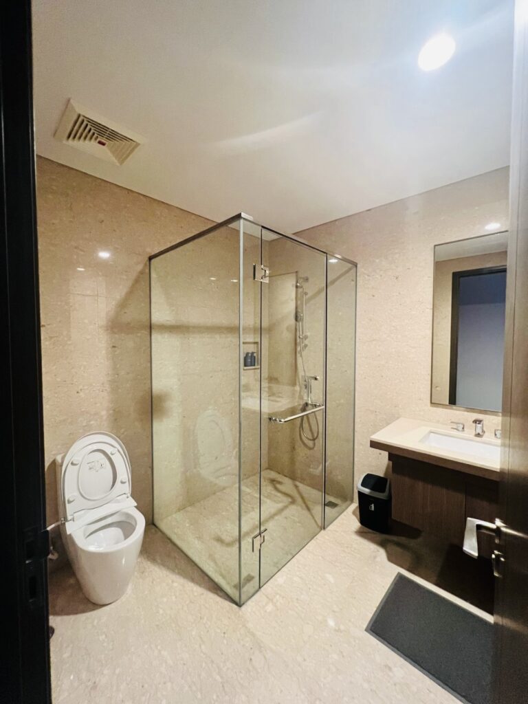 Kamar mandi modern di apartemen 31 Sudirman Suites Makassar dengan toilet, bilik shower kaca, dan wastafel, bagian dari unit 3 kamar + 1.