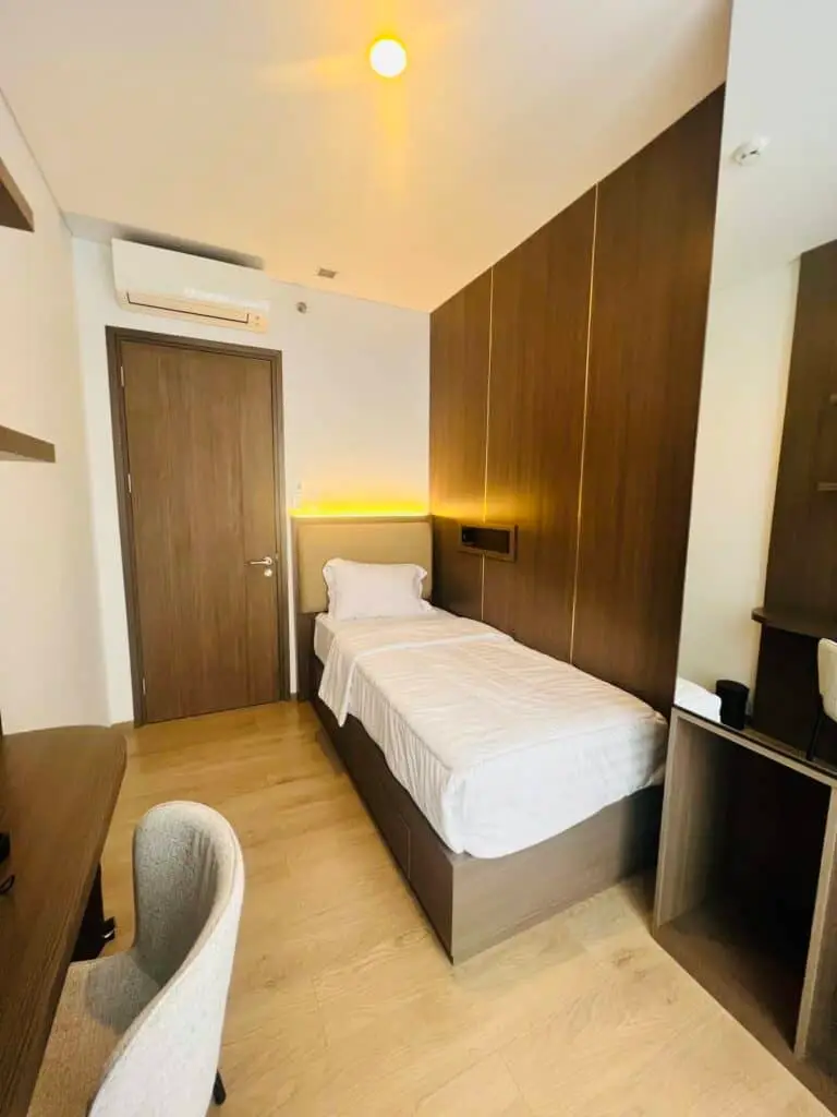 Kamar tidur kedua di apartemen 31 Sudirman Suites Makassar dengan ranjang single, headboard berlampu, dan meja belajar.