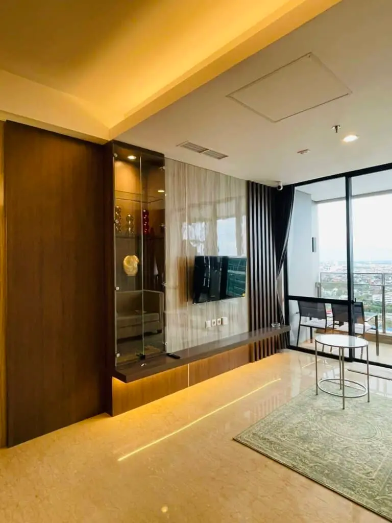 Ruang keluarga di apartemen 31 Sudirman Suites Makassar dengan panel TV modern, lemari pajangan kaca, dan pemandangan kota dari balkon.