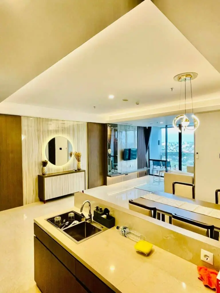 Pemandangan dari dapur modern ke arah ruang makan dan ruang tamu di apartemen 31 Sudirman Suites Makassar, menunjukkan konsep interior terbuka.